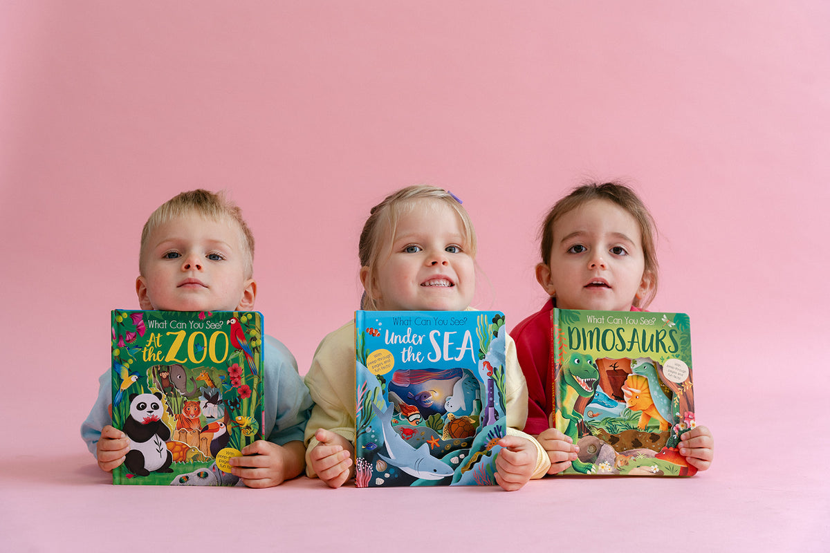 Three young children sitting side by side, each holding a colorful children's picture book against a solid pink background. The books are titled 'What Can You See? At the Zoo', 'Under the Sea', and 'What Can You See? Dinosaurs'.