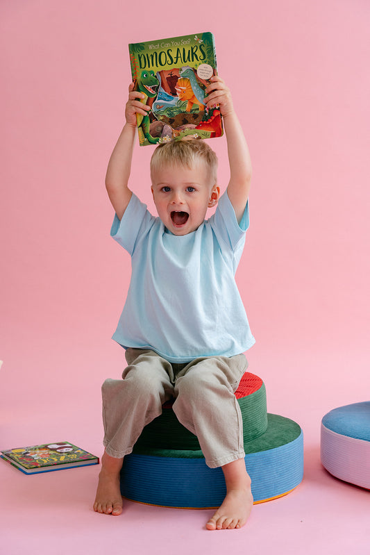 A young child wearing a light blue t-shirt and beige pants sits on colorful round foam blocks, holding a 'Dinosaurs' book above their head against a solid pink background.