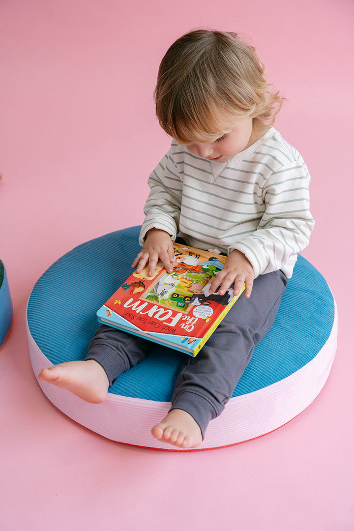 A toddler sits barefoot on a round blue and pink cushion, holding a colorful children's book against a solid pink background.